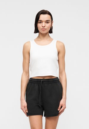 Vrouw met kort donker haar, gouden oorringen, een wit cropped tanktop en zwarte shorts met koordje, staand tegen een effen achtergrond.