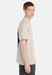 Μπεζ βαμβακερό T-shirt με στρογγυλή λαιμόκοψη, κοντά μανίκια και πολύχρωμο γραφικό σχέδιο στην πλάτη με περίπλοκα μοτίβα και διακοσμητικά στοιχεία.