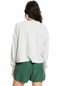 Eine Frau mit langen, dunklen Haaren trägt ein lockeres, hellgrau-weiß gestreiftes Langarmoberteil und grüne Shorts, steht mit dem Rücken zur Kamera.