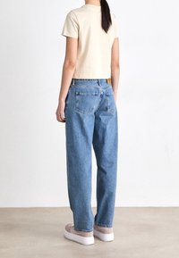 Donna vestita con una maglietta beige chiaro a maniche corte e jeans blu larghi, in piedi su un pavimento beige contro una parete bianca.