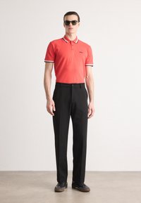 Rød polo skjorte med kontrasterende svarte og hvite striper på kragen og mansjettene, kombinert med svarte bukser og svarte sko, modell med solbriller.