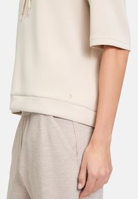 Jasno beżowa bluzka z krótkim rękawem o luźnym kroju, gładkiej teksturze i subtelnym metalicznym akcentem u dołu, w zestawie z miękkimi beżowymi spodniami.