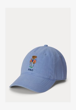 Jasnoniebieska czapka z daszkiem z wyhaftowanym misiem w okularach przeciwsłonecznych, kolorowej kurtce, dżinsach i trampkach, nad tekstem "POLO" na przednim panelu.