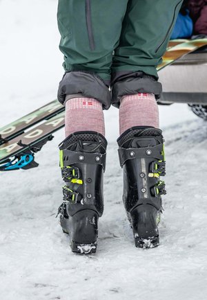 Persoon met zwarte skischoenen met neon groene gespen, roze sokken met een rood kruisdetail en groene broek staand op sneeuw bij ski's.