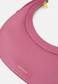 Różowa skórzana torebka o textured surface, zaokrąglonym kształcie, złotym zamku błyskawicznym i subtelnym logo wytłoczonym z przodu.