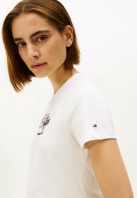 Hvid bomulds t-shirt med korte ærmer, der har et burgunder broderet logo på forsiden og et lille logo på ærmet.