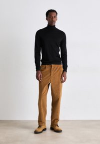 Pull en col roulé noir, texture côtelée; pantalon en velours côtelé marron à coupe droite; chaussures beige avec lacets noirs; fond neutre.