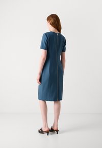 Blue fitted dress with short sleeves, back zipper closure, and a knee-length hem. Smooth texture. Brown mules with curved heels.