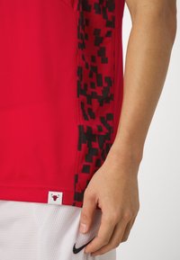 Camiseta deportiva roja con una tela de malla texturizada, que presenta un diseño pixelado negro en el costado y una etiqueta de logo en el dobladillo.