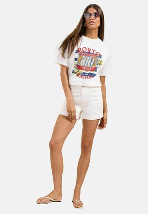 Naine kannab valget graafilise kujutisega T-särki, millel on kiri "Porto Portugal" ja kalamotiiv, valgeid lühikesi pükse, päikeseprille ja beeže sandaale, seisab valgel taustal.