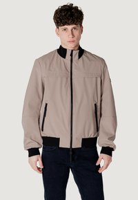 Giacca bomber beige con colletto alto, chiusura frontale con zip e polsini neri texturizzati. Presenta due tasche laterali con zip.