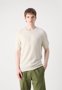 Maglietta a maniche corte color crema a coste con scollatura rotonda. Indossata con pantaloni verdi oliva a vestibilità rilassata, che evidenziano la texture e la silhouette.