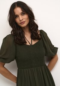 Rochie verde măsliniu cu un corset smocat, mâneci bufante și o fustă fluidă. Prezentă curele subțiri și un material texturat.