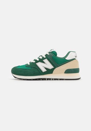 Grüner und beiger New Balance Sneaker mit weißen Schnürsenkeln und Logo, Mesh- und Veloursleder-Obermaterial, auf weißem Hintergrund.