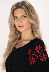 Schwarze Bluse mit Puffärmeln, die mit roten geometrischen Stickereien an den Schultern versehen ist. Weicher Stoff, offener Ausschnitt und Kordelzug-Detail.