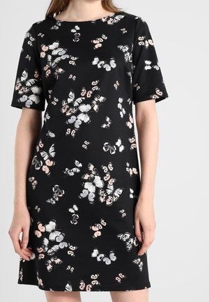 Femme portant une robe noire à manches courtes avec des motifs de papillons blancs et rose pâle sur un fond uni.