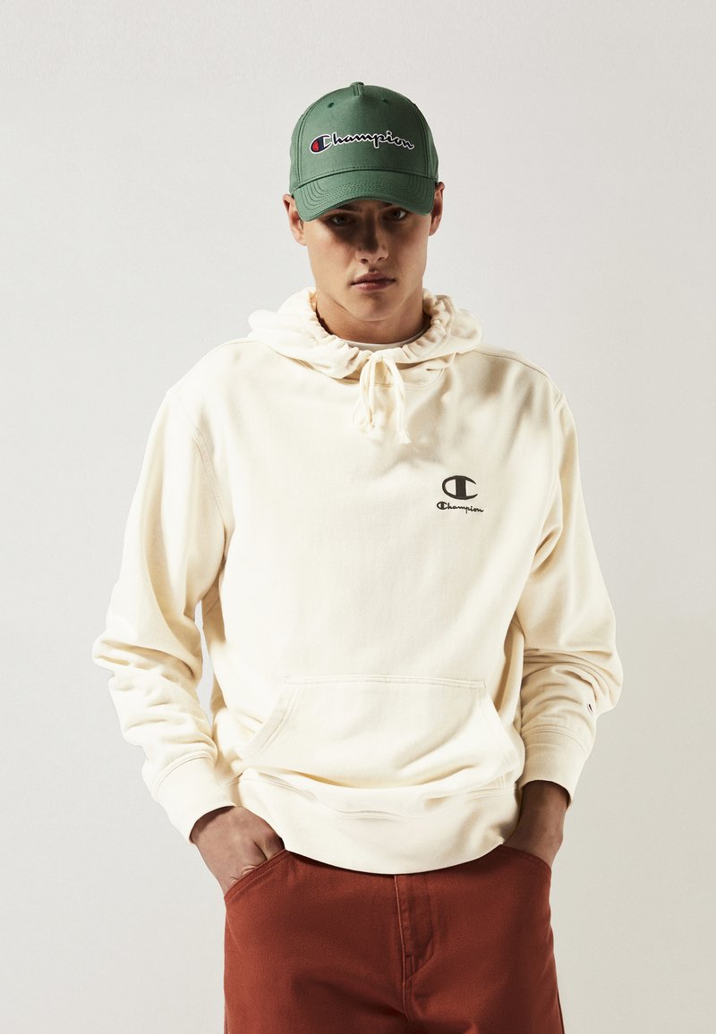 Jeune homme portant un sweat à capuche beige de la marque Champion, une casquette verte Champion et un pantalon rouille, les mains dans les poches, devant un fond uni.