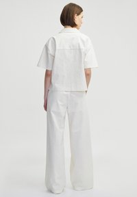 Camisa blanca de lino de manga corta con un detalle de yugo en la parte posterior, combinada con pantalones holgados de pierna ancha. Diseño minimalista y líneas limpias.