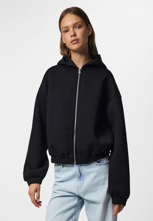 Ung kvinde iført en sort lynlåshoodie med hætte og lyseblå jeans, stående mod en ensfarvet hvid baggrund.