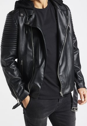 Mann trägt schwarze gesteppte Leder-Bikerjacke mit silbernen Reißverschlüssen, schwarzen Hoodie, schwarzes T-Shirt und schwarze Jeans, Hände in den Taschen.