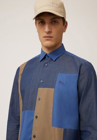 Button-up shirt featuring blue and brown patchwork design, cotton material, long sleeves, pointed collar, and left chest logo embroidery.