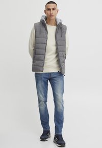 Grå puffervest med hette over en lys genser, kombinert med blå jeans og svarte joggesko. Glatt tekstur med quilted design.