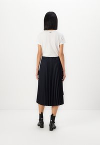 Tricou alb cu mânecă scurtă, cu detaliu logo pe spate, purtat cu o fustă midi plisată bleumarin și cizme negre până la gleznă. Textură a țesăturii netedă.