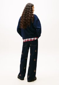 Μπλε φούτερ, πουκάμισο καρό από κάτω και σκούρο τζιν παντελόνι με πολύχρωμη κέντημα. Μακριά, κυματιστά μαλλιά και χοντρά μαύρα παπούτσια.