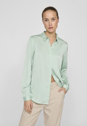 Bluză din satin verde deschis, cu guler, mânecă lungă, deschidere cu nasturi și detaliu strâns pe umeri, asortată cu pantaloni bej.