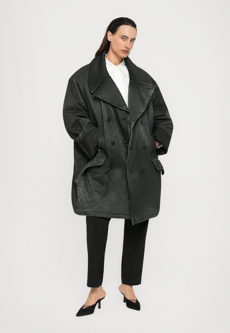 Femme aux cheveux foncés portant un manteau noir oversize à double boutonnage, une chemise blanche à col, un pantalon noir ajusté et des mules à talons noirs sur fond blanc.
