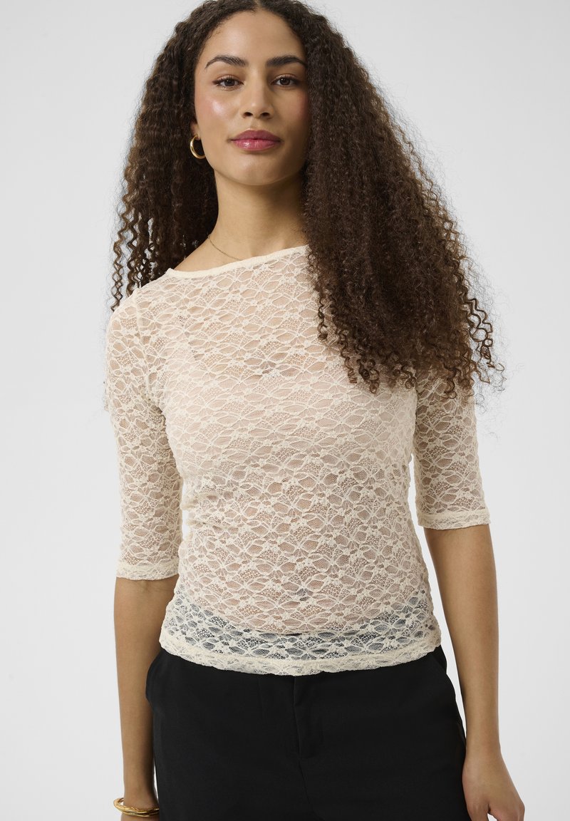 Femme aux longs cheveux bouclés porte un haut en dentelle blanche transparente à manches trois-quarts et un pantalon noir sur fond uni.