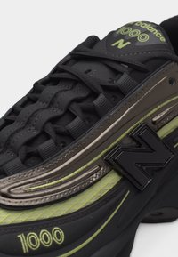 Basket New Balance 1000 noire et verte avec des lacets noirs, un grand logo noir "N" et des détails métalliques sur le côté.