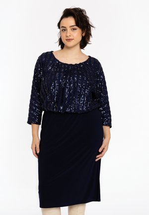 Kvinde iført en navyblå paliettop med trekvartlange ærmer og en matchende ensfarvet midinederdel, stående mod en hvid baggrund.