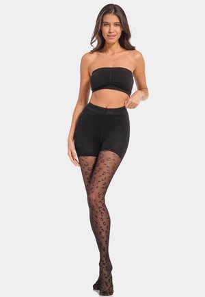 Femme portant un haut bandeau noir et un short taille haute avec des collants transparents noirs à imprimé léopard, debout devant un fond uni.