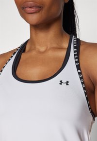 Top blanco sin mangas con detalles en negro, que presenta un escote redondeado y tirantes que exhiben la marca "Under Armour". Textura de tela suave.
