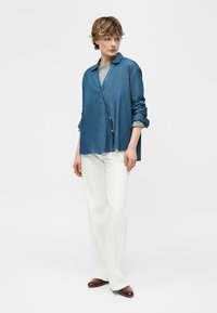 Mujer con cabello corto que lleva una camisa azul de mezclilla sobre una camiseta gris, pantalones blancos de pierna ancha y zapatos marrones, de pie con las manos relajadas.