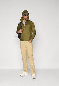 Lightweight olive green jacket with a hood, gray shirt, beige pants, white sneakers, and a black backpack. Sunglasses complete the outfit.