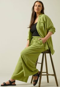 Groene linnen shirt en bijpassende wijde broek, gecombineerd met zwarte sandalen. De outfit heeft een relaxte pasvorm en een elastische tailleband.