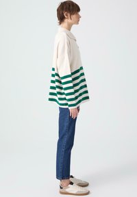 Breiclied in crème met groene horizontale strepen, voorzien van een kraag en een losse pasvorm. Gepaard met donkerblauwe jeans en casual sneakers.