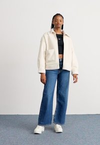 Giacca in pile oversize color crema con colletto e tasca frontale, abbinata a un top corto nero e jeans a gamba larga blu. Sneakers bianche.