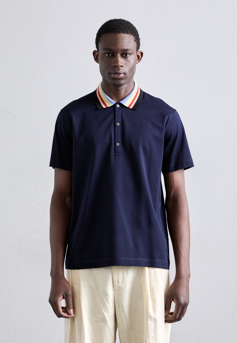 Polo shirt bleu marine avec un col rayé rouge, jaune et noir. Il présente une coupe classique et un rabat de trois boutons.