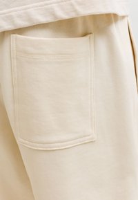 Gros plan d’un pantalon en tissu beige montrant une poche arrière carrée avec des coutures visibles sur le côté droit.