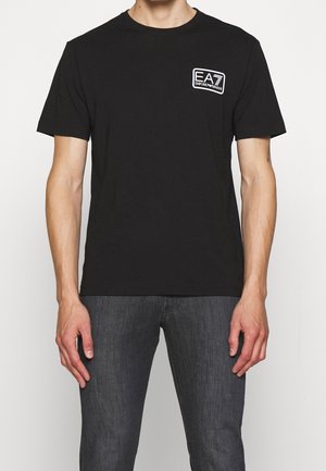 Mężczyzna w czarnej koszulce z krótkim rękawem z małym białym logo "EA7 Emporio Armani" na lewej części klatki piersiowej, w połączeniu z ciemnoszarymi dżinsami.