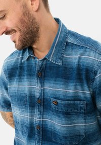 Knapp-up skjorte lavet af blå denim med vandrette hvide striber, en brystlomme og brune knapper. Har et blødt, tekstureret stof.