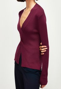 Pull en maille bordeaux avec un col en V profond, une coupe ajustée, des manches longues, des fentes latérales et une texture côtelée. Associé à un pantalon foncé.