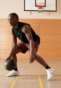Camisa sin mangas negra, pantalones cortos burdeos y zapatillas blancas. El jugador sostiene un balón de baloncesto negro en un gimnasio con paredes de madera y un aro.