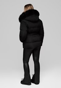 Černá péřová bunda s kapucí z umělé kožešiny, s páskem v pase a prošívaným designem. V kombinaci s černými zvonovými kalhotami a plochými černými botami.