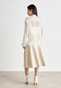 Weißes transparentes Bluse mit Rüscheldetails und zarter Stickerei. Kombiniert mit einem beigen Rock. Das Model trägt beige Stiefeletten.