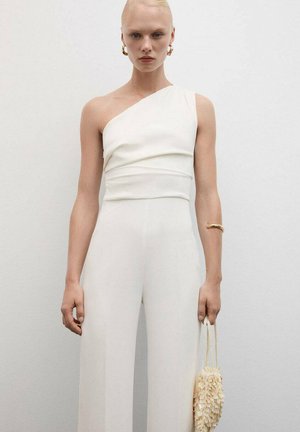 Witte, een-schouder jumpsuit met een taillering bodice en losse, brede benen; gecombineerd met een gestructureerde, met franjes versierde lichtgekleurde handtas.