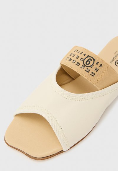 Μπεζ σανδάλι slide με ανοιχτά δάχτυλα, φαρδιά κρεμ λουράκι και ελαστική ταινία με αριθμό που δείχνει το μέγεθος 6, σε φωτεινό φόντο.